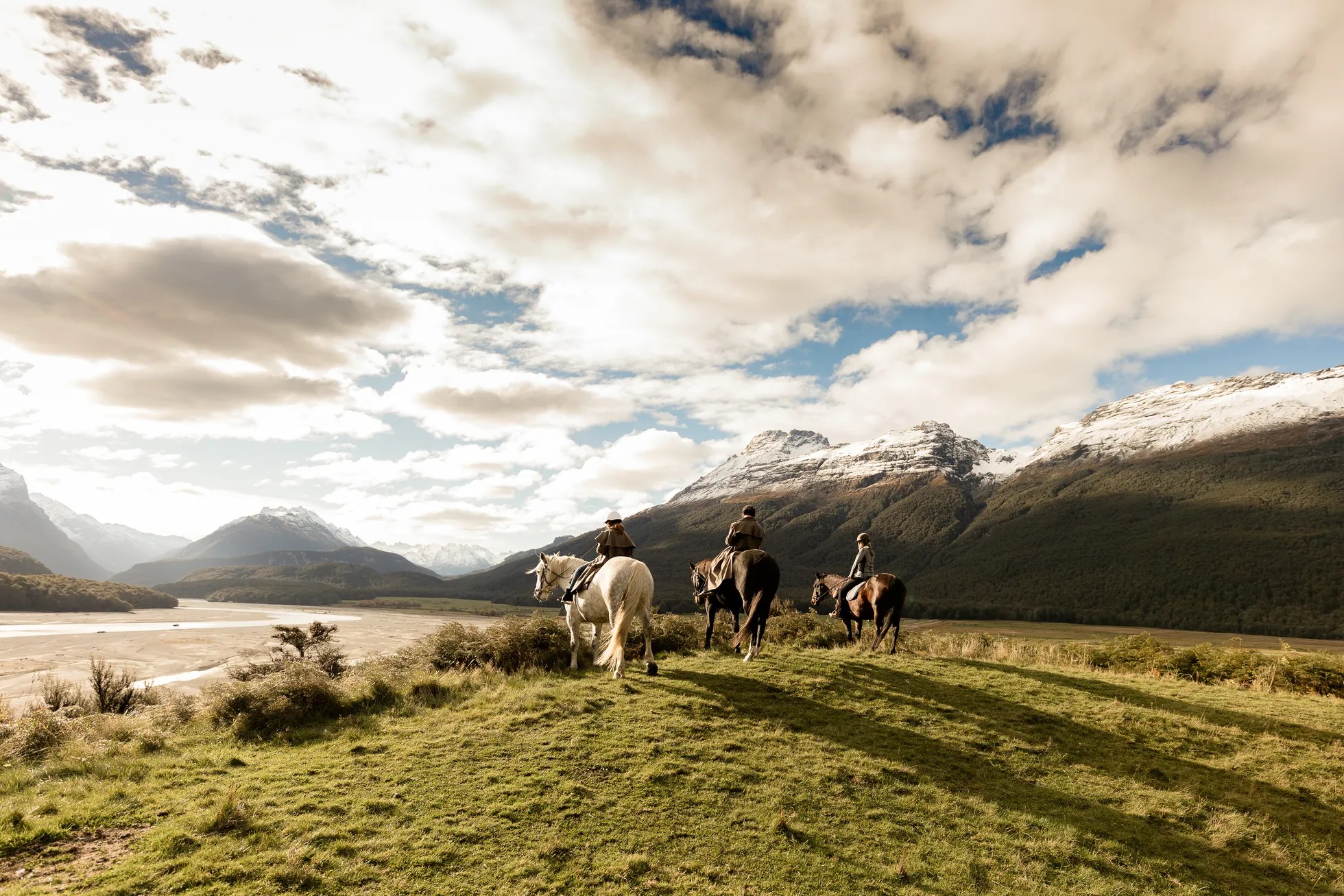 Three people riding horses in an a grassy field with mountains in the background.
