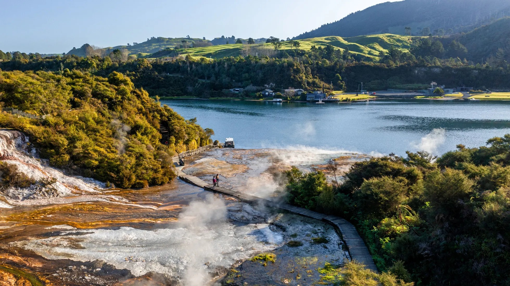 A geothermal terrace beside a lake. On top of the terrace is a wooden boardwalk, which two people are walking on.