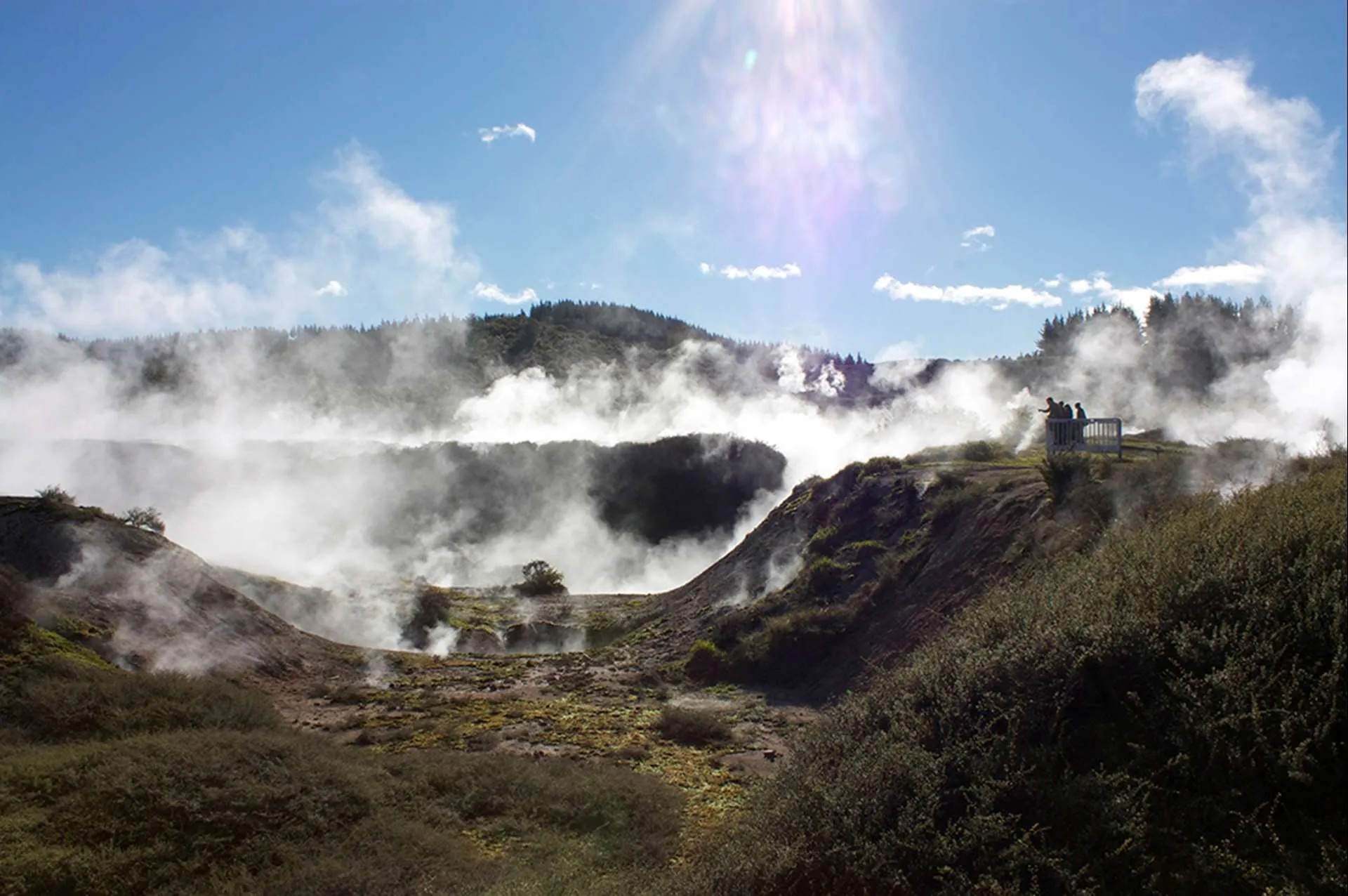 A hilly, crater-like environment filled with rising steam. A group of people stand at a lookout spot.