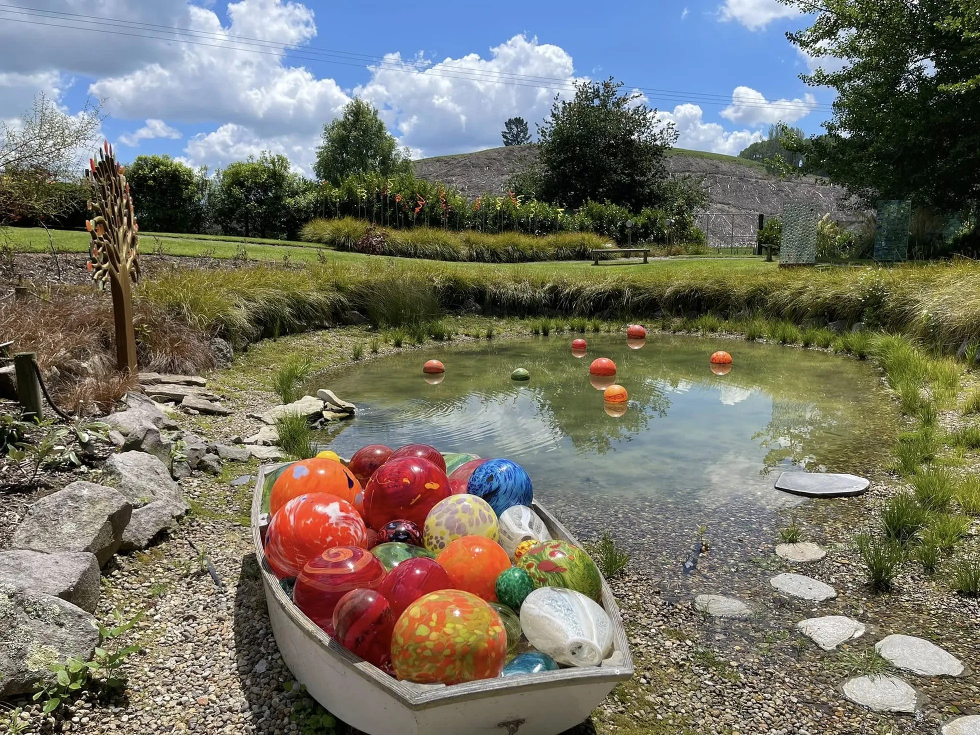 A boat on the edge of a small pond. The boat is filled with colourful glass orbs, and a few orbs are sitting on the water.