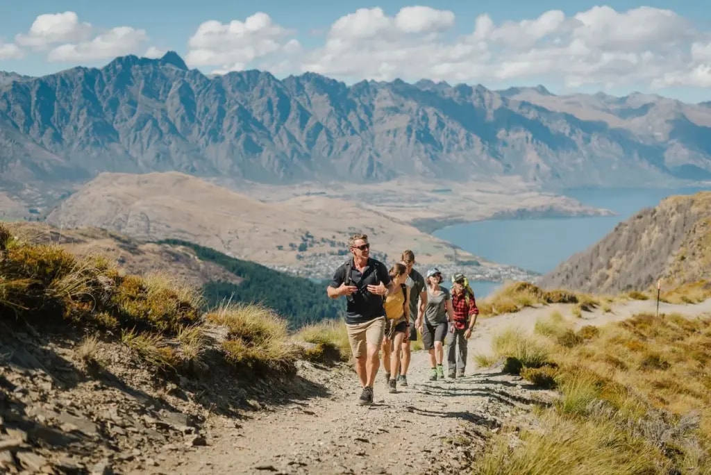 Man leading a group of walkers up a gravel mountain path, views of Queenstown and Lake Wakatipu afar.