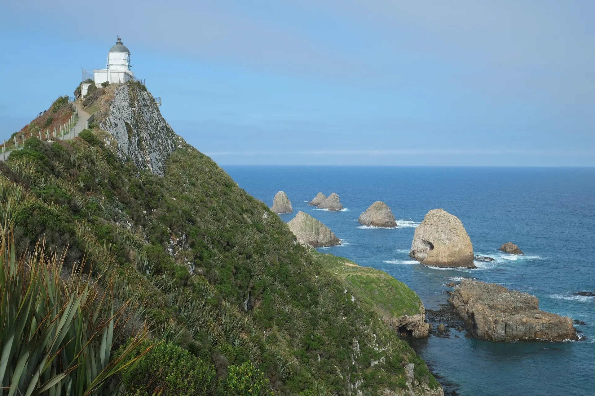 A cliff with a white lighthouse at the end. The surrounding waters have a few small islets