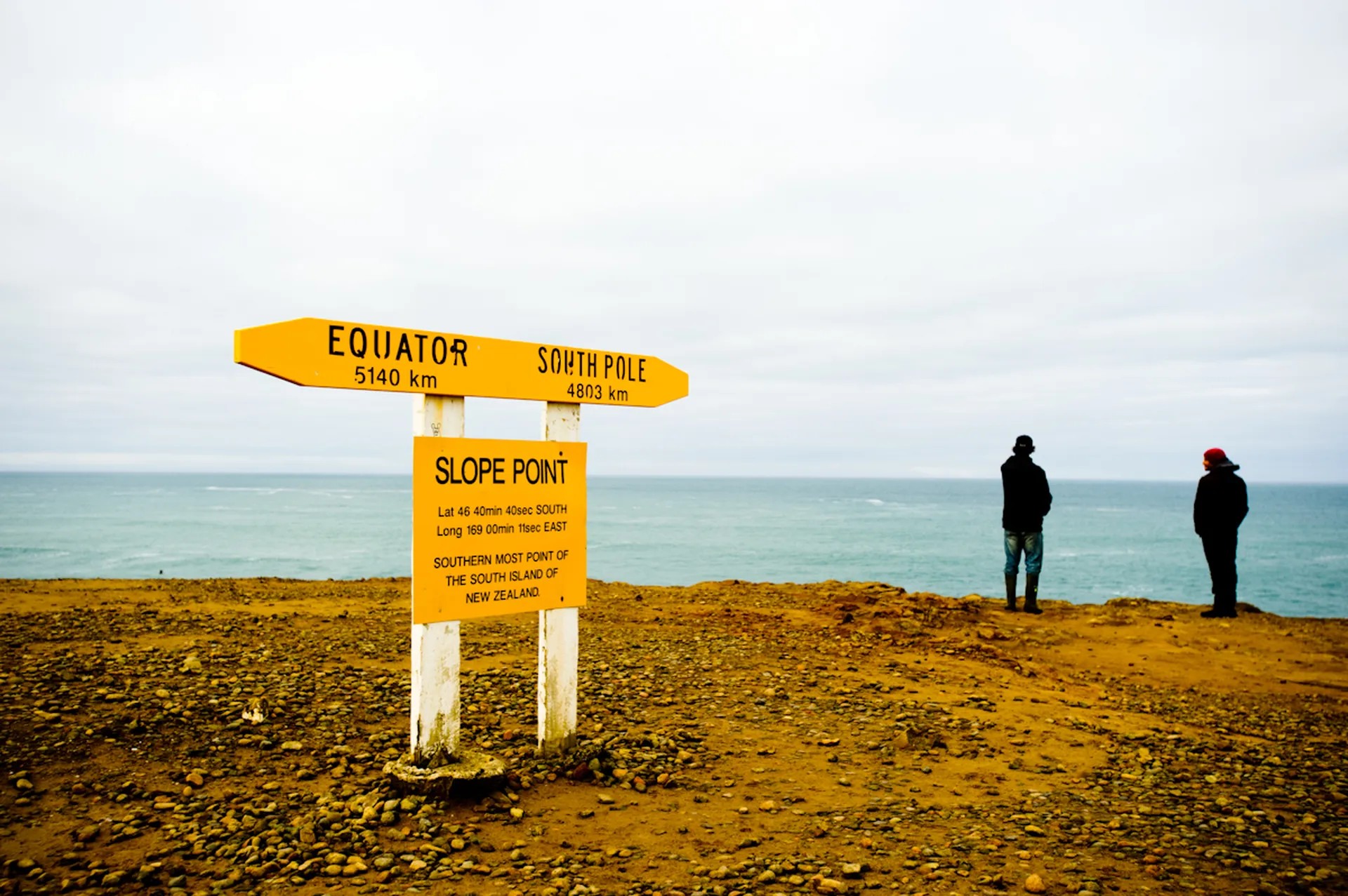 A cliff with a yellow sign that reads "Slope Point - Southernmost Point of the South Island of New Zealand" and points to the Equator and South Pole with distances. Two people stand in the back, looking at the sea