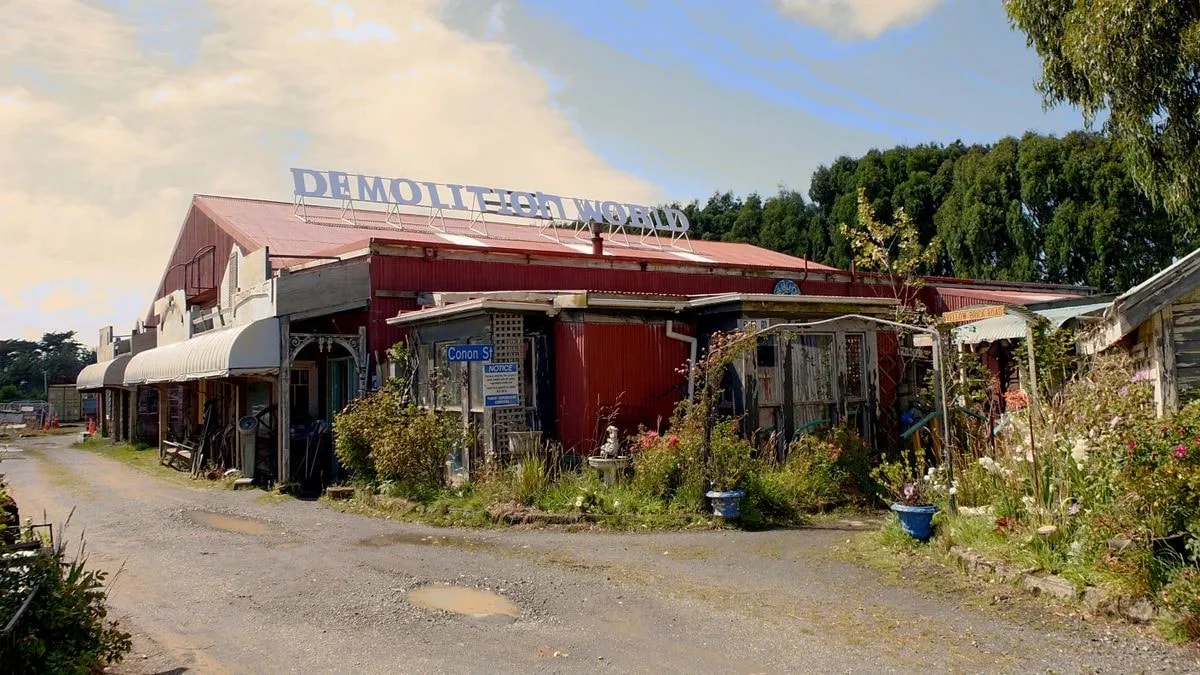 A shed-barn with a sign reading "Demolition World" on top; nearby are various sheds with flowers and objects beneath
